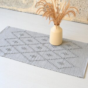 Puede incluir: Una alfombra tejida gris con un patr&oacute;n geom&eacute;trico de diamantes. La alfombra est&aacute; sobre un suelo blanco y un jarr&oacute;n con hierba seca est&aacute; en el fondo.