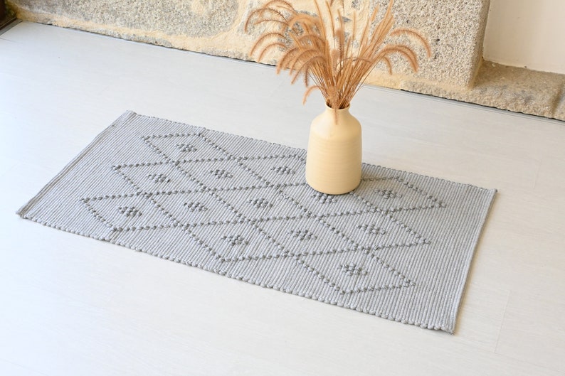 Puede incluir: Una alfombra tejida gris con un patr&oacute;n geom&eacute;trico de diamantes. La alfombra est&aacute; sobre un suelo blanco y un jarr&oacute;n con hierba seca est&aacute; en el fondo.
