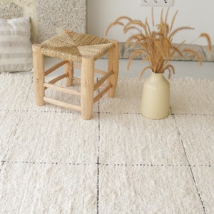 May include: A white woven rug with black stitching, a wooden stool with a woven seat, and a vase with dried grass.