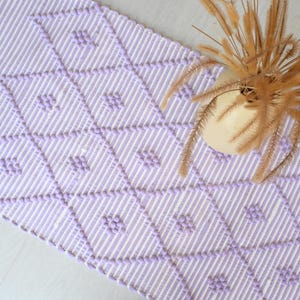 Könnte beinhalten: Lavendel-weiß gewebter Tischläufer mit Rautenmuster. Der Läufer hat einen gestreiften Hintergrund mit erhabenen Rautenformen. Eine kleine Vase mit getrockneten Pflanzen steht auf der Ecke des Läufers.