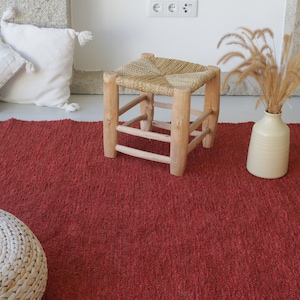 May include: A red woven rug with a white woven stool and a vase with dried grass on top of it.