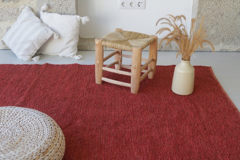 May include: A red woven rug with a white woven stool and a vase with dried grass on top of it.