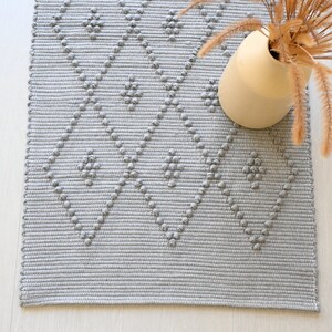 Puede incluir: Una alfombra tejida gris y blanca con un patr&oacute;n de diamante. La alfombra tiene una superficie texturizada y est&aacute; hecha de fibras naturales.