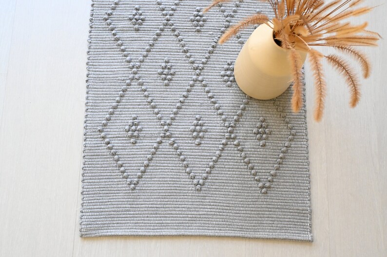 Puede incluir: Una alfombra tejida gris y blanca con un patr&oacute;n de diamante. La alfombra tiene una superficie texturizada y est&aacute; hecha de fibras naturales.