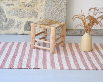 Può includere: Un piccolo sgabello in legno naturale con seduta intrecciata è appoggiato su un tappeto a righe rosa e bianche. Un vaso beige contiene erbe secche. Il tappeto è lungo circa 91 cm. La scena è ambientata contro un muro di pietra.