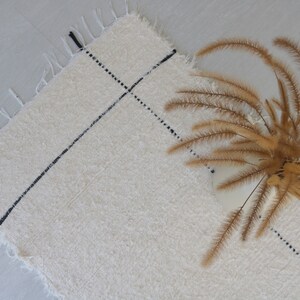 May include: A white rug with black lines woven into the fabric. The rug has a fringe border and is lying flat on a light gray surface. A bouquet of dried brown grasses is lying on the rug.