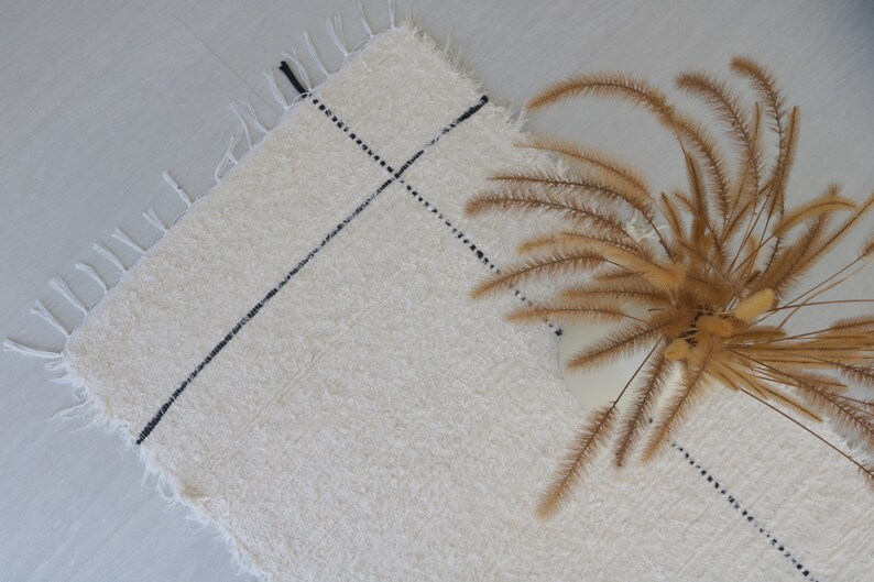 May include: A white rug with black lines woven into the fabric. The rug has a fringe border and is lying flat on a light gray surface. A bouquet of dried brown grasses is lying on the rug.