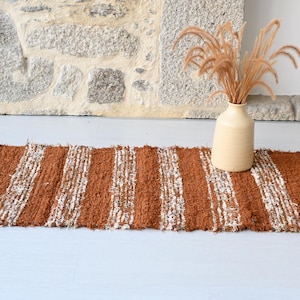 Puede incluir: Una alfombra rectangular tejida a mano con rayas verticales marrones y blancas. Un jarrón beige con plantas secas se encuentra sobre la alfombra. La alfombra tiene un borde con flecos.