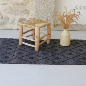 May include: A black and white patterned rug with a woven texture. A small wooden stool with a woven seat sits on the rug. A vase with dried grass sits on the floor next to the rug.