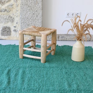 May include: A green woven rug with a natural wood stool with a woven seat. The stool is in the centre of the rug. A vase with dried grass is on the right side of the rug.