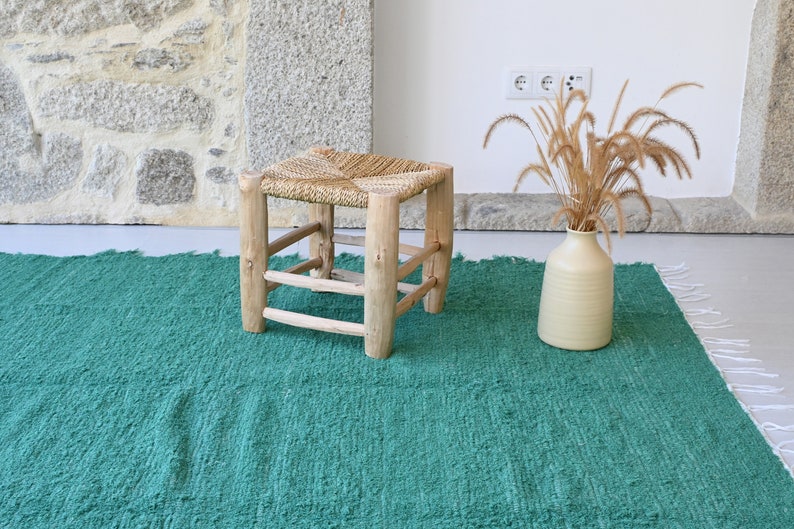 May include: A green woven rug with a natural wood stool with a woven seat. The stool is in the centre of the rug. A vase with dried grass is on the right side of the rug.