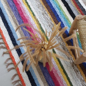 Puede incluir: Una alfombra de rayas coloridas con flecos en los bordes, un taburete de madera con asiento tejido y un arreglo de flores secas.