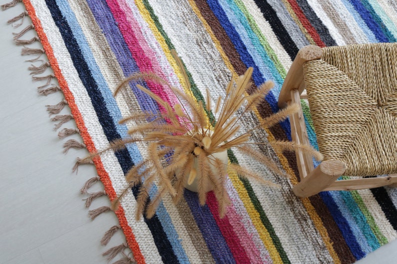 Puede incluir: Una alfombra de rayas coloridas con flecos en los bordes, un taburete de madera con asiento tejido y un arreglo de flores secas.