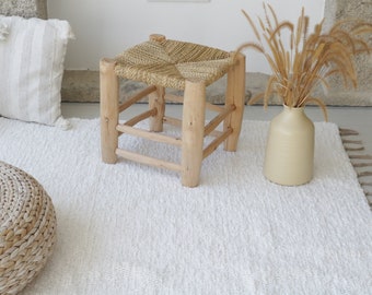 May include: A white fluffy rug with a woven straw stool and a round woven ottoman. A vase with dried grass is on the rug.