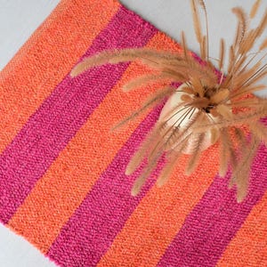 Op de afbeelding: Een rechthoekig tapijt met afwisselende verticale strepen in fel oranje en felroze. Een kleine gouden vaas met gedroogd siergras staat op het tapijt. Het tapijt is gemaakt van een textuurrijk materiaal.