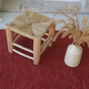 May include: A red woven rug with a natural woven stool and a vase of dried grass.