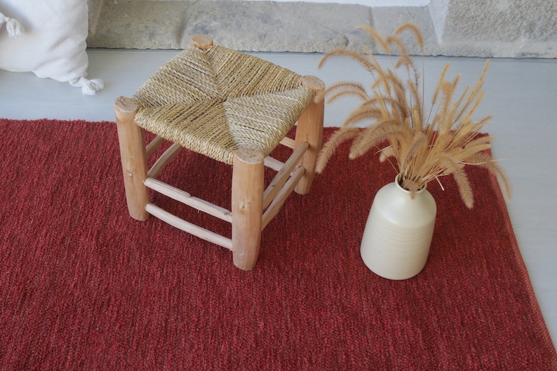May include: A red woven rug with a natural woven stool and a vase of dried grass.