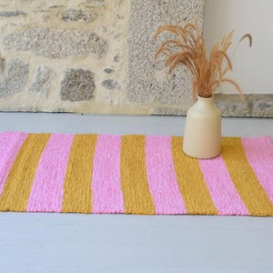 May include: A rectangular area rug featuring alternating vertical stripes in bright pink and mustard yellow. The rug has a textured, woven appearance and lies flat. A light beige ceramic vase containing dried grasses sits on the rug's surface.