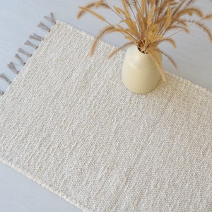 Puede incluir: Una alfombra tejida de color crema con flecos en los bordes. La alfombra est&aacute; colocada sobre una superficie gris claro con un jarr&oacute;n de flores secas encima.