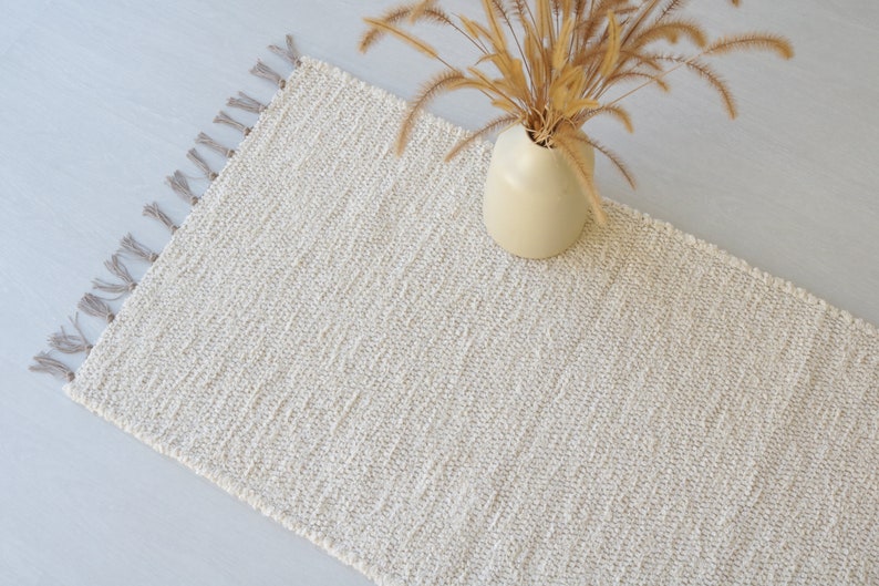 Puede incluir: Una alfombra tejida de color crema con flecos en los bordes. La alfombra est&aacute; colocada sobre una superficie gris claro con un jarr&oacute;n de flores secas encima.