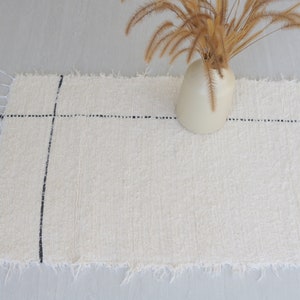May include: A white woven rug with black lines, featuring a frayed edge. The rug is placed on a light gray floor with a vase of dried grass in the background.