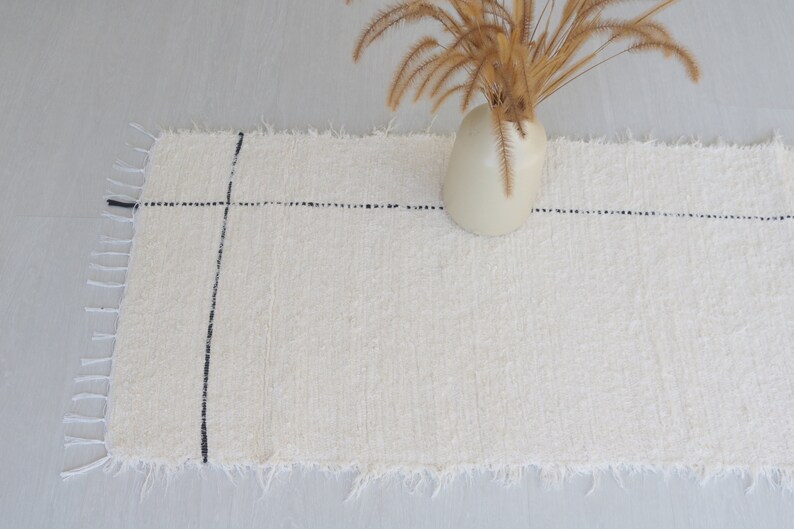 May include: A white woven rug with black lines, featuring a frayed edge. The rug is placed on a light gray floor with a vase of dried grass in the background.