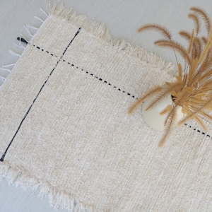 Peut inclure: Un tapis blanc avec des lignes noires et un bord effiloché. Un vase avec de l'herbe sèche est sur le tapis.
