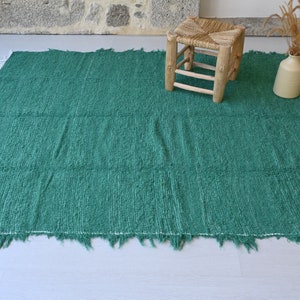 May include: A green woven rug with a fringed edge. The rug is on a light wood floor and there is a small wooden stool and a vase with dried flowers in the background.
