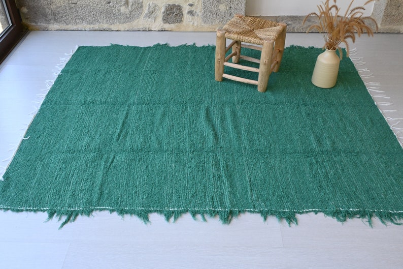 May include: A green woven rug with a fringed edge. The rug is on a light wood floor and there is a small wooden stool and a vase with dried flowers in the background.