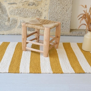 Può includere: Un tappeto rettangolare caratterizzato da strisce verticali alternate color giallo senape e bianco sporco. Il tappeto presenta una trama tessuta e strutturata. Uno sgabello in legno a quattro gambe con seduta in paglia intrecciata.
