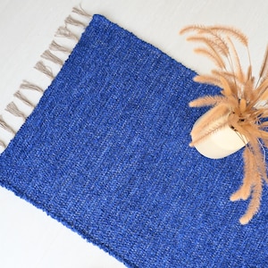 Può includere: Un tappeto blu tessuto con frange sui bordi. Il tappeto è realizzato in un materiale morbido e testurizzato e ha una leggera lucentezza. Un arrangiamento di fiori secchi è posizionato sul tappeto.