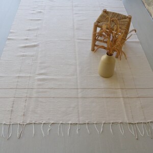 Puede incluir: Una alfombra blanca tejida con un borde de flecos. Un peque&ntilde;o taburete de madera con un asiento tejido y un jarr&oacute;n con flores secas est&aacute;n sobre la alfombra.