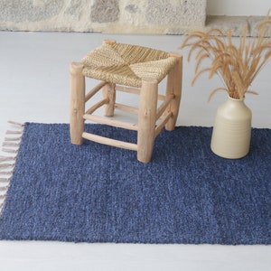 Puede incluir: Una alfombra azul tejida con flecos en los bordes y un taburete de madera natural con un asiento tejido. La alfombra está sobre un suelo blanco y hay un jarrón con hierba seca en el fondo.