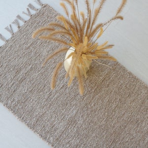 May include: A beige woven rug with fringe on the edges. The rug is laying flat on a white surface with a vase of dried grass in the center.