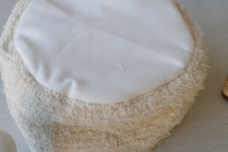 May include: A round, cream-colored pouf with a fluffy, shaggy texture. The pouf has a smooth, white fabric top.