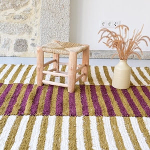 May include: A striped rug with white, olive green, and purple stripes. A small wooden stool with a woven seat sits on the rug. A cream-coloured vase holds dried plants.