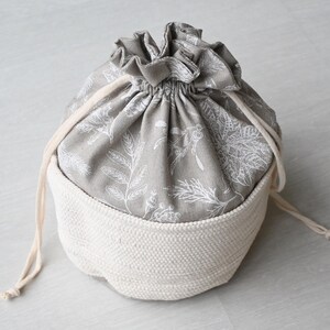 Pu&ograve; includere: Una borsa con coulisse grigia e bianca con una base bianca intrecciata. La parte superiore della borsa &egrave; in tessuto grigio con un motivo floreale bianco. Il cordoncino &egrave; color crema.
