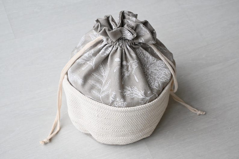Pu&ograve; includere: Una borsa con coulisse grigia e bianca con una base bianca intrecciata. La parte superiore della borsa &egrave; in tessuto grigio con un motivo floreale bianco. Il cordoncino &egrave; color crema.