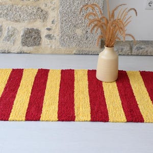 Peut inclure: Tapis rectangulaire à rayures verticales rouges et jaunes. Un vase beige avec des plantes séchées est posé sur le côté droit. Le tapis est fait d'une matière texturée. Dimensions : environ 120 cm de long et 60 cm de large.
