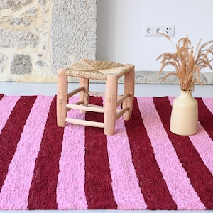 May include: A pink and burgundy striped rug with a wooden stool with a woven straw seat in the centre.