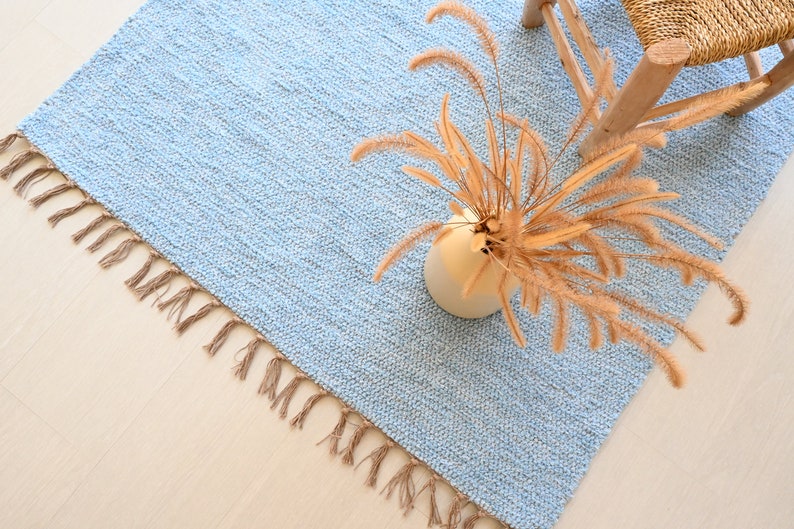 Pu&ograve; includere: Un tappeto blu chiaro con un bordo a frangia e un vaso di erba della pampa secca in cima.