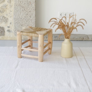 Puede incluir: Una alfombra tejida blanca con un taburete tejido natural y un jarr&oacute;n con hierba seca.
