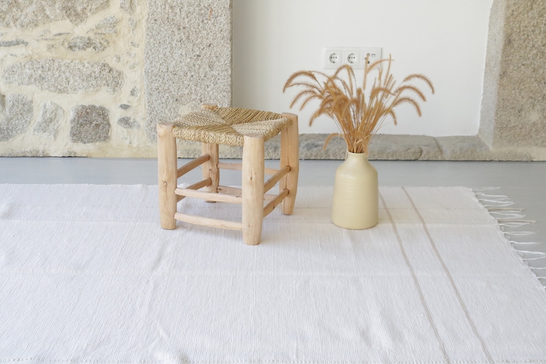 Puede incluir: Una alfombra tejida blanca con un taburete tejido natural y un jarr&oacute;n con hierba seca.