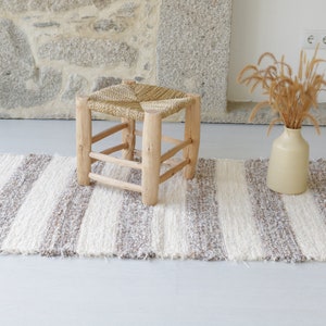 May include: A white and brown striped rug with a woven texture and frayed edges. A wooden stool with a woven seat sits on the rug.