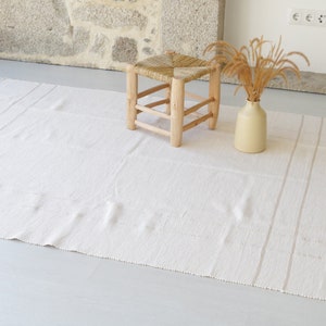 Puede incluir: Una alfombra blanca tejida con flecos en los bordes y dos sutiles rayas que recorren el centro. Un peque&ntilde;o taburete de madera con un asiento tejido se encuentra sobre la alfombra.