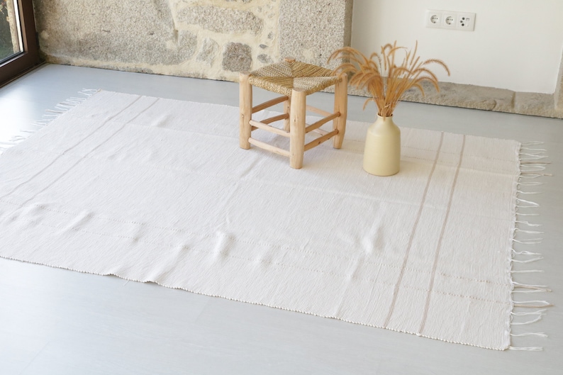 Puede incluir: Una alfombra blanca tejida con flecos en los bordes y dos sutiles rayas que recorren el centro. Un peque&ntilde;o taburete de madera con un asiento tejido se encuentra sobre la alfombra.
