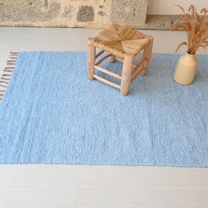 Pu&ograve; includere: Un tappeto blu chiaro con frange sui bordi. Il tappeto &egrave; su un pavimento bianco con uno sgabello di legno e un vaso con fiori secchi.