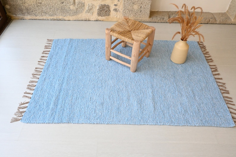 Pu&ograve; includere: Un tappeto blu chiaro con frange sui bordi. Il tappeto &egrave; su un pavimento bianco con uno sgabello di legno e un vaso con fiori secchi.