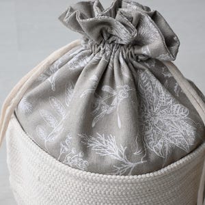 Pu&ograve; includere: Una borsa con coulisse con base in tessuto color crema e parte superiore a fantasia floreale grigia. La parte superiore &egrave; chiusa con una coulisse color crema. Il motivo floreale presenta disegni botanici bianchi.