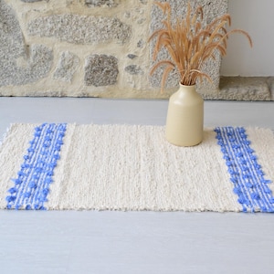 Puede incluir: Una alfombra rectangular tejida en color blanco roto con dos bandas verticales de pompones azules y flecos en los bordes cortos. Un jarrón de cerámica beige con tallos de trigo secos se encuentra sobre el lado derecho de la alfombra.